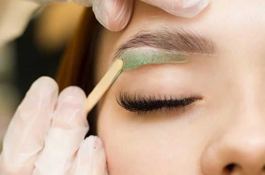 Eyebrow being waxed with green wax applied by a professional using a wooden stick.