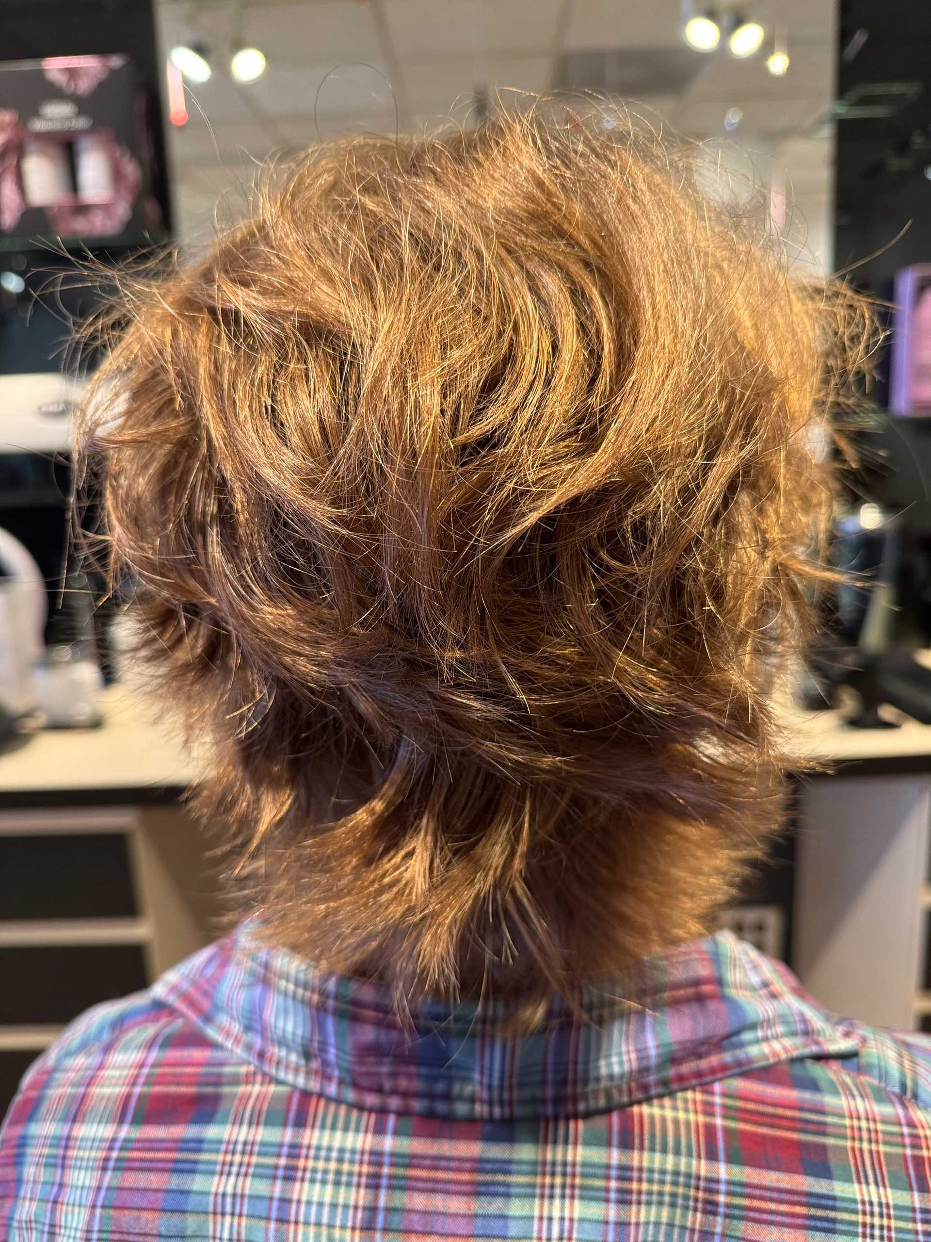Back view of a person with tousled, short brown hair wearing a plaid shirt in a salon.
